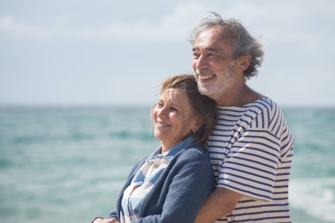 casal de idosos se abraçando em uma praia e sorrindo para o horizonte